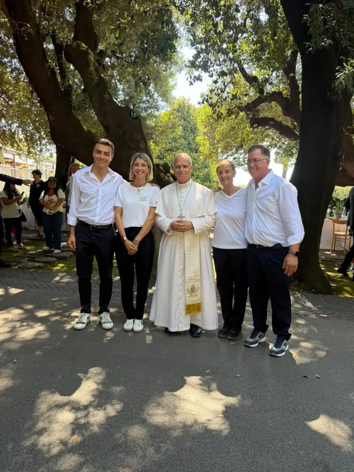 Five people pose beneath large trees in a garden, one in a white cassock